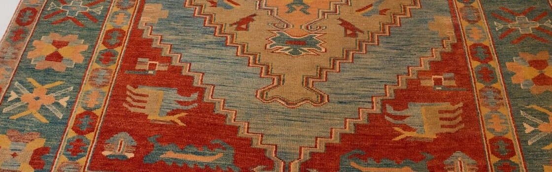 Nahaufnahme eines Teppichs mit gestuftem Medaillon in Sand und Hellblau auf rotem Grund, an den Seiten eine türkisfarbene Bordüre